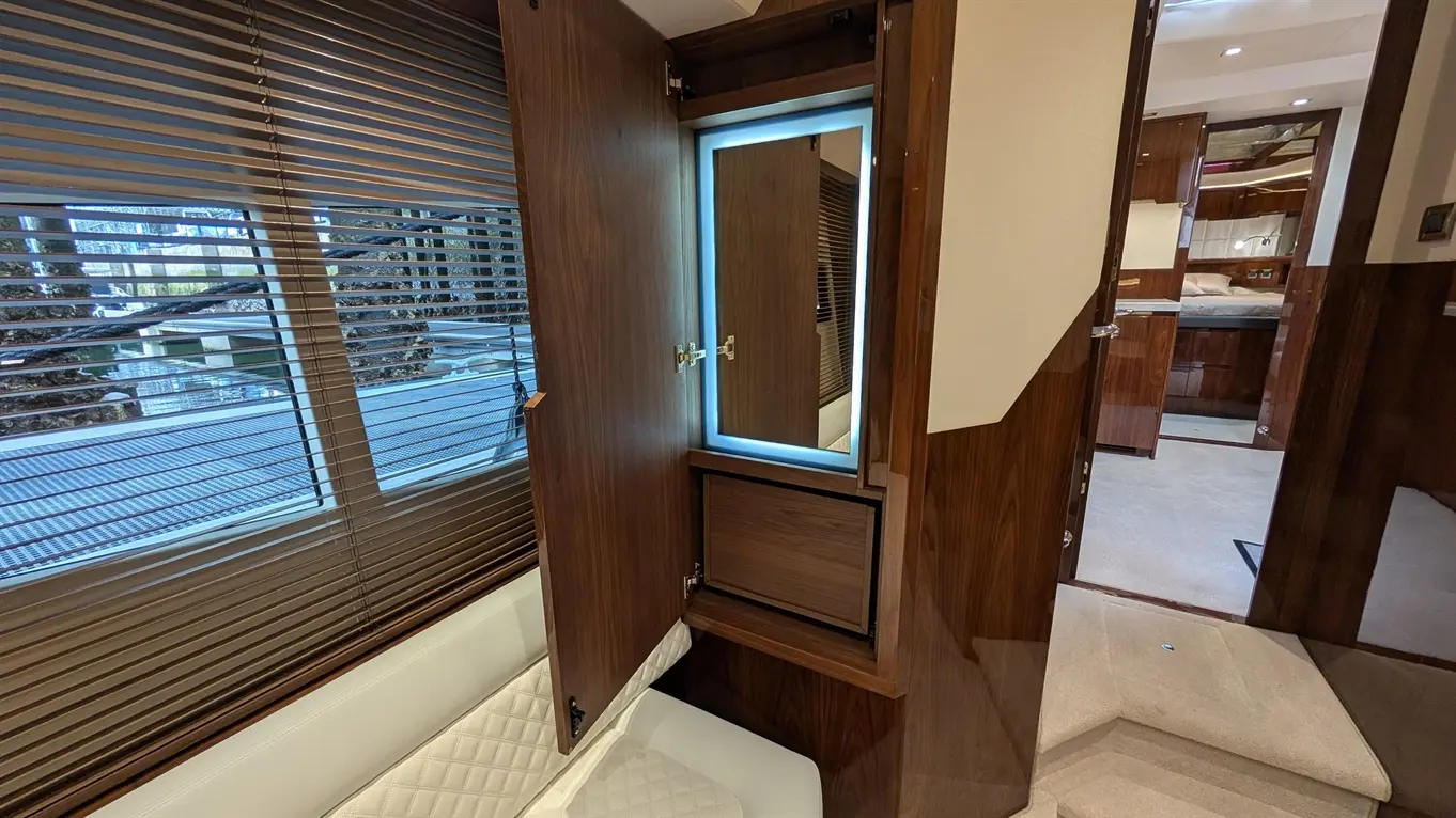 Interior of a luxury yacht showing a wooden cabinet with an illuminated mirror and a view through slatted blinds.
